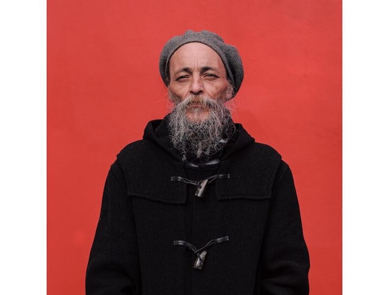 Keith Murrell - long grey beard in grey beret and black duffel coat against a block red background