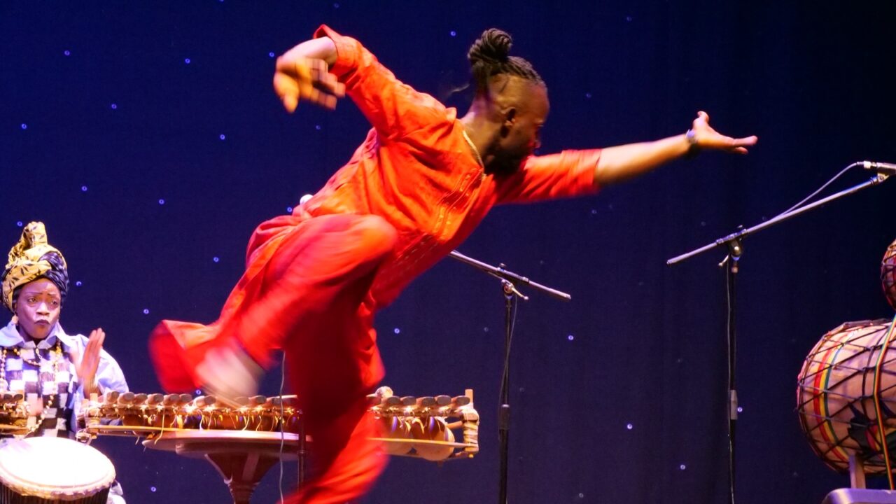 Salif Camara in red dancing on stage with Fatoumata Kouyate in the background playing the balafon