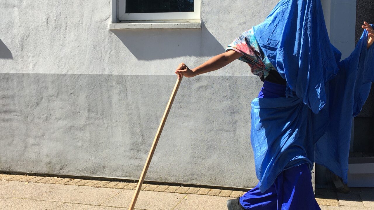 Person dressed in blue cloth, covering their head and face with walking stick, part of dance piece by BACA