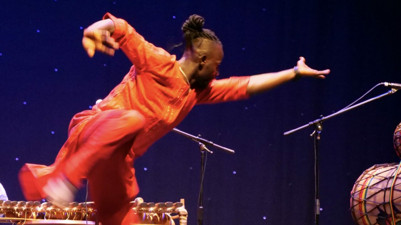 Salif leaping sideways in red costume dancing on stage at The Welfare, Ystradgynlais