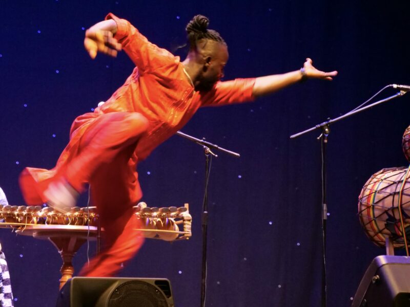 Salif leaping sideways in red costume dancing on stage at The Welfare, Ystradgynlais